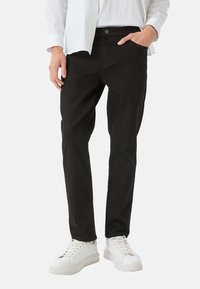 Jeans noirs en mélange de coton, coupe slim avec une fermeture à bouton, poches avant et texture lisse. Porté avec des baskets blanches.