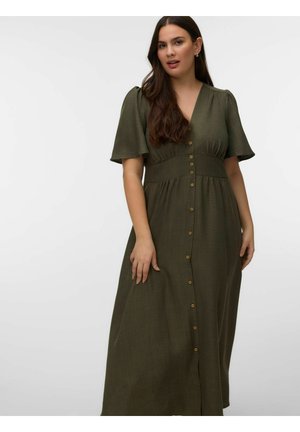 Kvinde med langt mørkt hår iført en lang olivengrøn kjole med knapper og korte pufærmer, stående foran en ensfarvet baggrund.