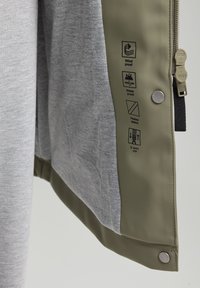 Harmaa kangas, jossa on sileä vedenpitävä oliivinvihreä kuori. Ominaisuuksiin kuuluvat tuulenpitävä muotoilu, lämpöeristetyt saumat, kaksisuuntainen vetoketju ja hoitosymbolit.