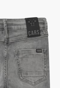 Harmaat farkut, joissa on musta nahkainen brändipatch vyötäröllä ja pieni brändilogo takataskussa. Kulunut pinta.