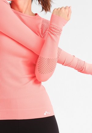 Kvinde der strækker armene ud iført en lyserød, tekstureret træningstop med lange ærmer og sorte leggings mod en ensfarvet hvid baggrund.