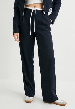 Pantalons évasés bleu marine en tissu doux, dotés d'une taille à cordon blanc et de détails plissés, associés à des baskets blanches.