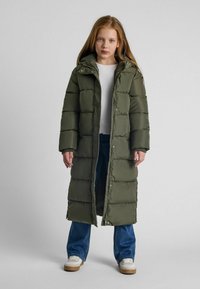 Garšs, polsterēts parka mētelis olīvzaļā krāsā ar kapuci, priekšējiem pogām un quilted tekstūru. Valkāts virs baltas krekla un ziliem džinsiem.