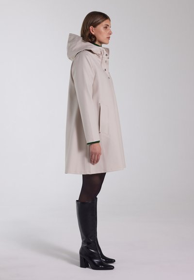 Beige regenjas met capuchon, knopen, zijzak, zachte textuur; gecombineerd met zwart kniehoog hakken. Zijaanzicht.