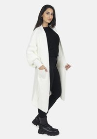 Cardigan long ouvert en tricot crème avec une texture côtelée, comportant de grandes poches et des manches tombantes, associé à un haut et un pantalon noir.