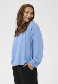 Top texturat de culoare albastru deschis, cu mâneci lungi, manșete elastice, croi lejer și guler rotund, asociat cu pantaloni negri.