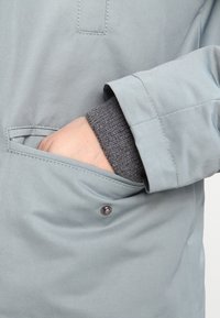 Main dans la poche latérale d'une veste gris clair avec un bord-côte gris foncé visible au poignet.