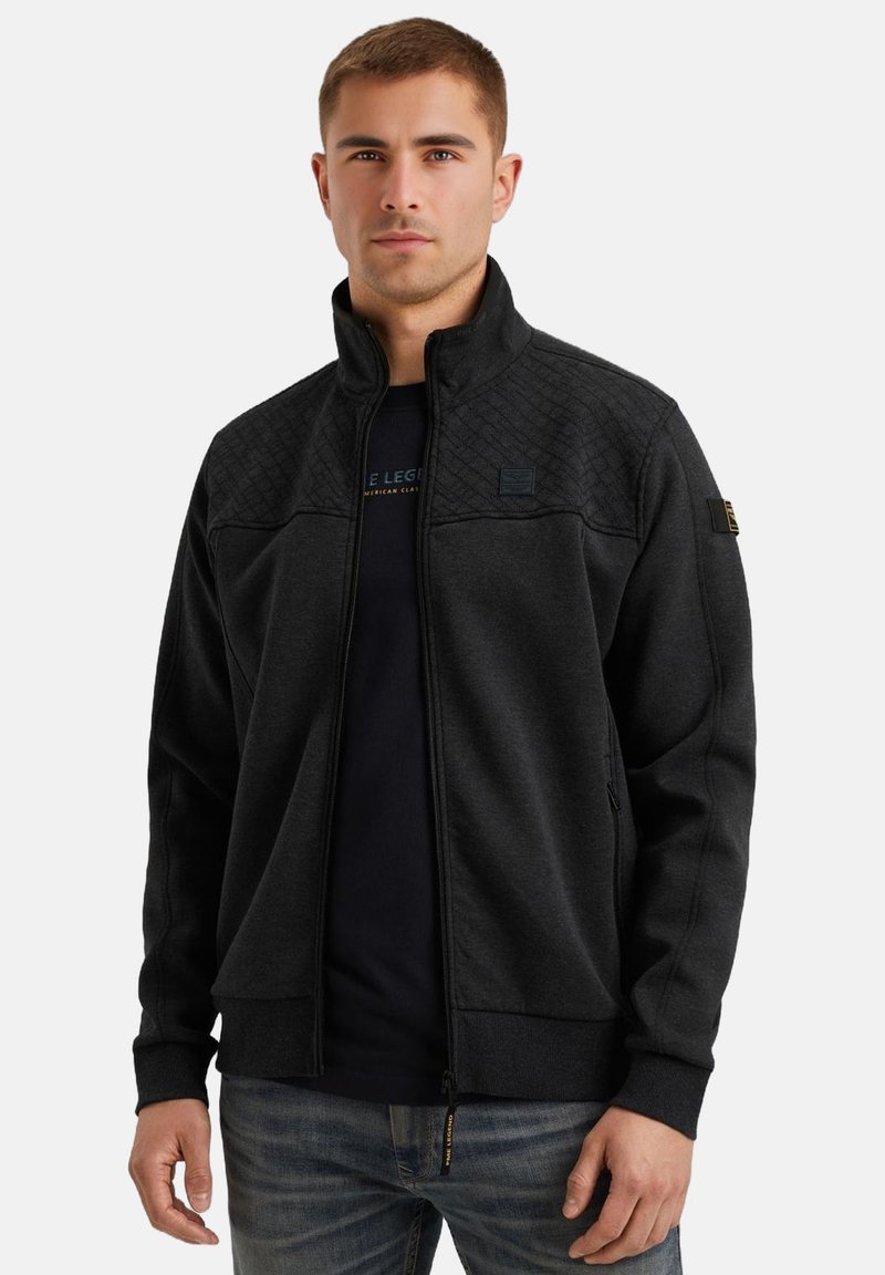 Jeune homme portant une veste matelassée sombre zippée sur une chemise noire et un jean bleu, se tenant devant un fond blanc uni.