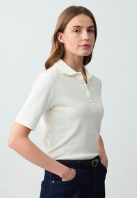Korte mouwen, crèmekleurig poloshirt met een gestructureerd ribbelpatroon, drie knoopsluitingen en een zachte kraag. Gepaarde met donkerblauwe jeans.