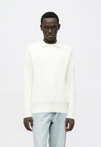 Polo de malha branca de manga comprida com gola e três botões. Combinado com jeans azul claro, apresentando um corte descontraído.