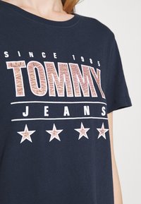 Námořnická modrá bavlněná tričko se zvýšeným nápisem "TOMMY" v texturované hnědé barvě, se bílými akcenty a pěti bílými hvězdami pod názvem značky.