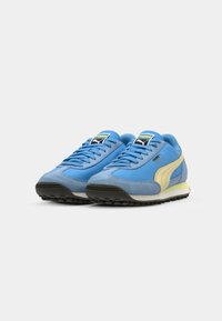 Sneakers azul em camurça e malha com sola bege, sola de borracha preta e etiqueta verde no língua. Apresenta atacadores e listra lateral em amarelo.