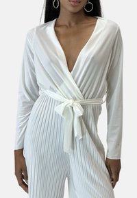 Mono blanco de manga larga con escote en V profundo, cintura con lazo y textura acanalada, fabricado en tela suave y con detalle decorativo de nudo.