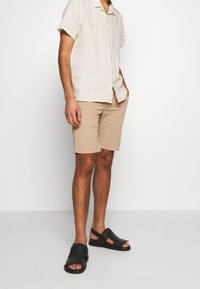 Pantaloni bermuda beige e una camicia in lino chiaro a maniche corte, abbinati a sandali neri in pelle con frange e punte aperte.