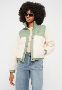 Fliisist jope rohelise ülemise ja kreemika alumise osaga, trukknappidega, soonikkoes varrukasäärtega ja väikese logoga rinnal.