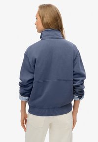 Sudadera azul con cuello alto, puños acanalados y un corte relajado. Presenta un panel de contraste en la parte posterior y una textura suave.