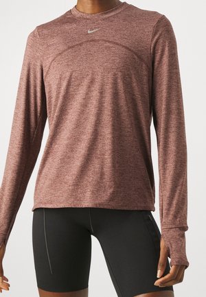 Persoon draagt een bruin gemêleerd sportshirt met lange mouwen en duimgaten, gecombineerd met zwarte nauwsluitende fietsbroekjes.