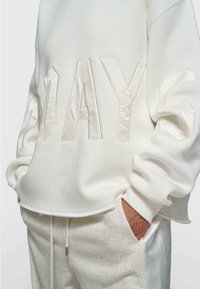 Sudadera blanca con la palabra "DAYS" en letras de tela en relieve, con un corte relajado y puños acanalados. Combinada con joggers de color claro.