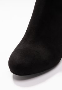 Zapato de tacón alto de ante negro que presenta una puntera redondeada y una textura suave. El tacón es sutil, realzando su diseño aerodinámico.