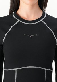 Chemise noire à manches longues avec détails de couture blanche, dotée d'un col rond et du logo "TOMMY JEANS" sur la poitrine.