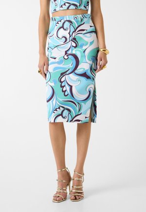 Donna con gonna a vita alta, lunghezza al ginocchio, con vortici astratti blu e bianchi, abbinata a sandali dorati con tacco alto e cinturini.