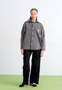 Camicia in denim grigia con abbottonatura e dettaglio tasca, abbinata a pantaloni larghi neri e sandali neri a scivolo su un pavimento verde.