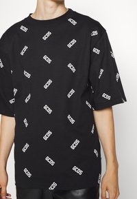 Camiseta de algodón negra con un patrón repetido del logo "GCOS" en blanco, de mangas cortas y con un diseño clásico de cuello redondo.