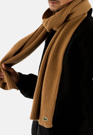 Bufanda de punto marrón con textura acanalada, hecha de un material suave, que presenta un pequeño logo verde. Colgada alrededor del cuello, sostenida en la mano contra un fondo negro.