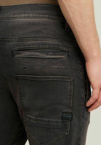 Κοντινό πλάνο ενός χεριού ανθρώπου που ακουμπάει στο πλάι, φορώντας σκούρο γκρι τζιν με πολλές πίσω τσέπες και ετικέτα G-Star Raw.