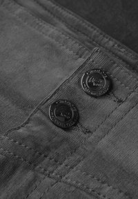 Un tissu gris au toucher doux présente deux boutons métalliques noirs sur une poche, embossés avec "Nous aimons." Des coutures sont visibles le long des bords.
