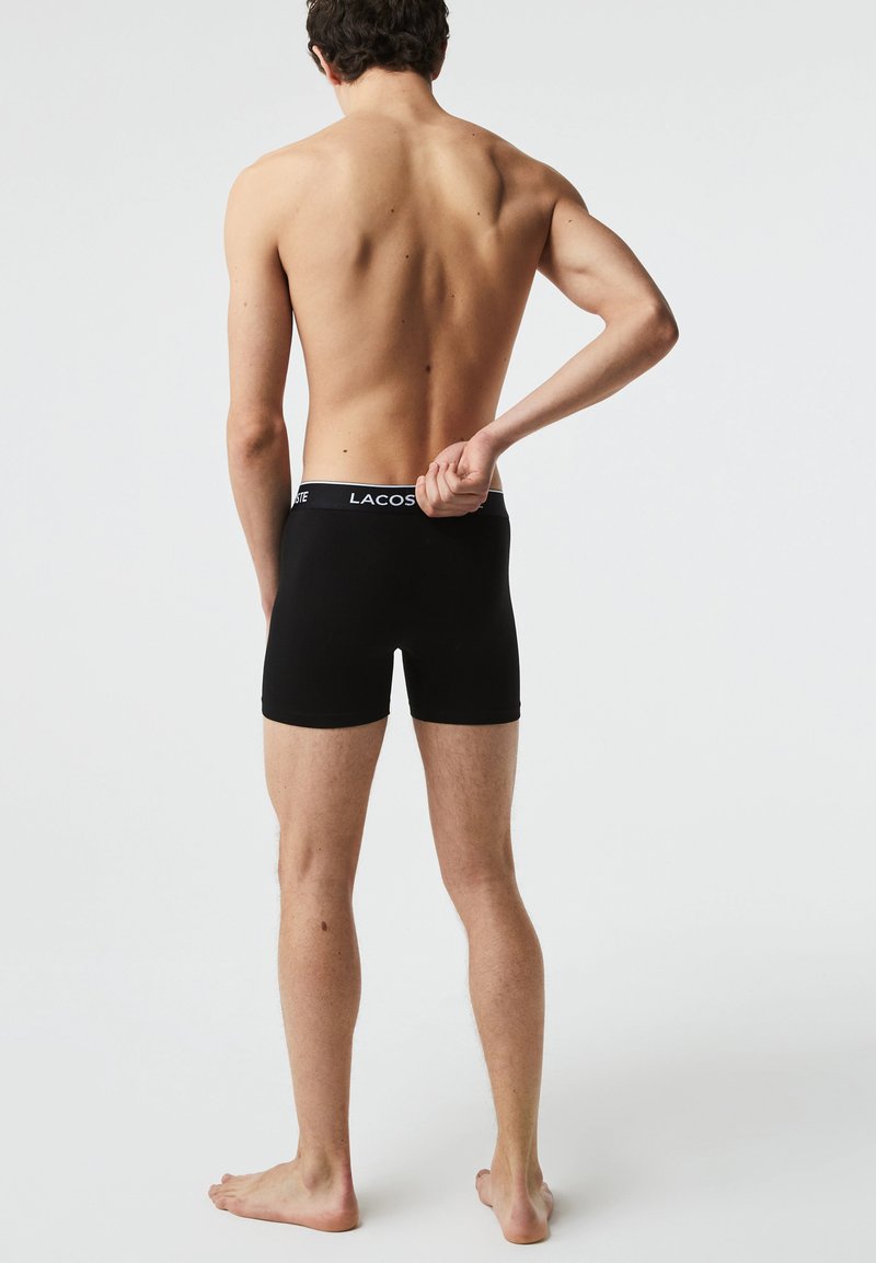 Schwarze Boxer-Shorts mit einem weißen Bund, auf dem das "LACOSTE"-Logo zu sehen ist. Das Material wirkt glatt; der Schnitt ist enganliegend mit einem mittelhohen Design.