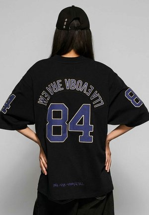 Persoon met lang donker haar, een zwarte pet en een oversized zwart T-shirt met het nummer 84 en gebogen tekst op de achterkant.