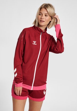 Frau, die ihre Haare richtet, trägt eine bordeauxrote und rosa hummel Sportjacke und passende Shorts und steht vor einem schlichten weißen Hintergrund.