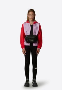 Färgbrodd vindjacka i rosa och rött, svart midjeväska, svarta leggings och grå skor med robusta sulor. Modellen står mot en vit bakgrund.