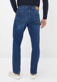 Jean bleu en denim avec cinq poches, design à coupe droite, coutures piquées et un patch en cuir beige à la taille. Texture lisse.