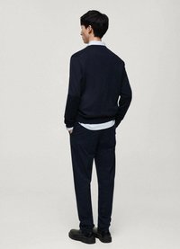 Marinblått stickat tröja med långa ärmar, parat med mörka byxor och en ljus skjorta under. Svarta skor fulländar outfiten.