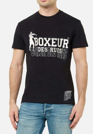 Mann trägt schwarzes T-Shirt mit weißem Text "Boxeur des Rues" und einer Boxer-Silhouette, kombiniert mit hellblauen Jeans.