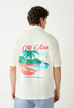 Man wearing a short-sleeve white shirt with a colorful "Cote d'Azur" beach scene and sailboat printed on the back.