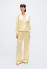 Γυναίκα που στέκεται ξυπόλητη φορώντας ένα σετ πιτζάμες από παλ κιτρινοσατέν ύφασμα με φαρδύ γιακά και μπλε λεπτομέρειες στο γιακά και στα τελειώματα του παντελονιού.