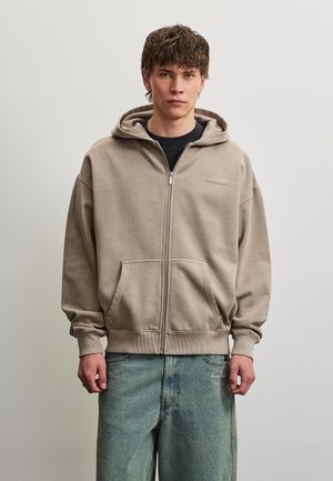 Jeune homme portant un sweat à capuche beige avec fermeture éclair, poches frontales et jean bleu clair, debout devant un fond gris clair uni.