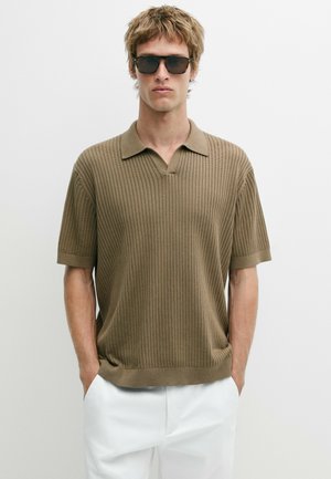 Mann trägt braunes, geripptes Poloshirt mit kurzen Ärmeln und weiße Hosen, die Hände in den Taschen, trägt auch dunkle Sonnenbrille, steht vor einfarbigem Hintergrund.