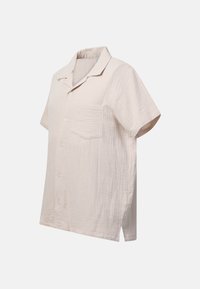 Chemise beige à manches courtes à boutons, en tissu texturé avec une poche poitrine devant, présentée sur un fond neutre.