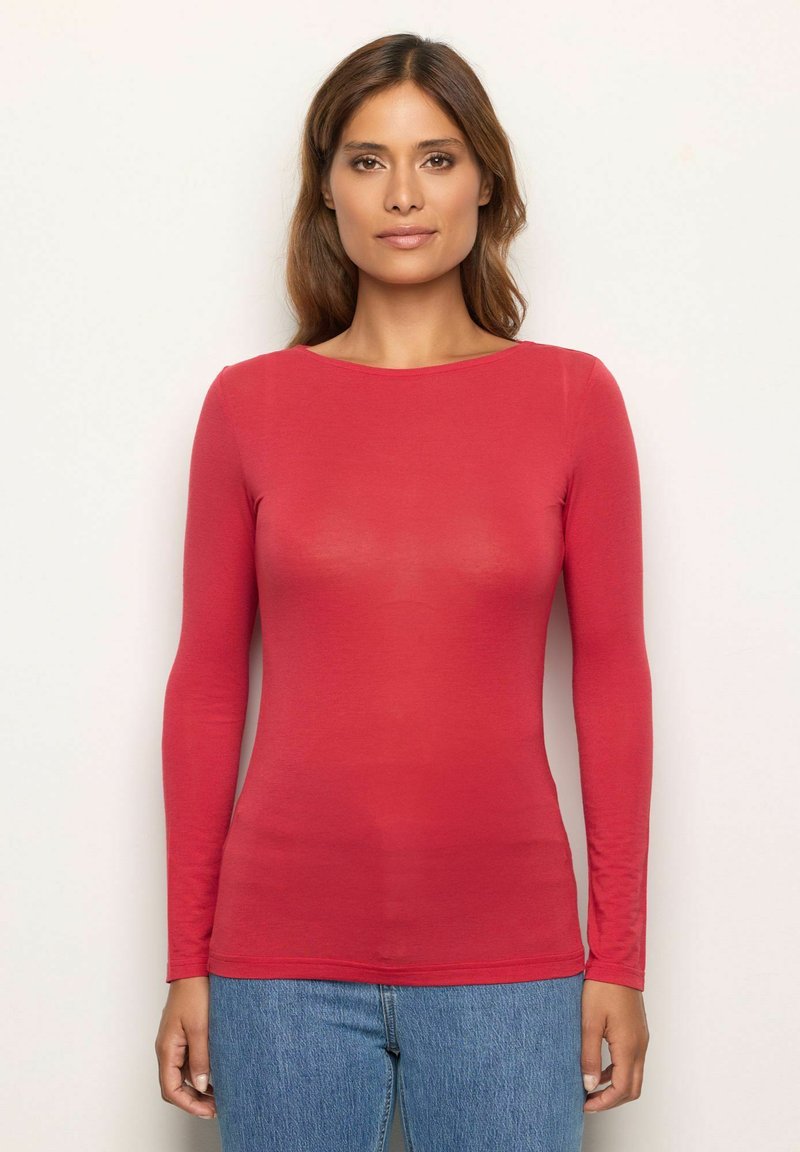 Mujer con cabello castaño, vistiendo una blusa roja de manga larga ajustada y jeans azules, de pie contra un fondo ligero y sencillo.