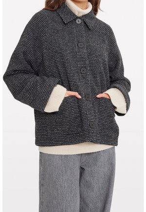 Personne portant une veste noire et blanche à carreaux boutonnée sur un pull crème et un pantalon gris, les mains dans les poches de la veste.