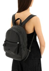 Mochila de cuero negro con acabado texturizado, forma redondeada, bolsillos frontales con doble cremallera, correas ajustables y un asa superior como acento.