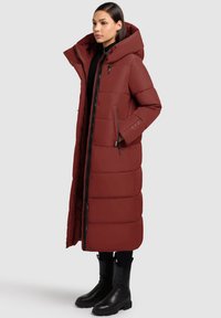 Ilga, pūsta tamsiai raudonos spalvos striukė su aukštu apykakliu ir kapišonu. Yra kišenės su užtrauktukais ir priekinis užtrauktuko uždarymas. Medžiaga atrodo atspari vandeniui.