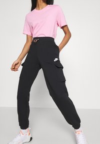 Černé cargo kalhoty Nike s elastickým pasem, dvěma bočními kapsami a bílým logem. V kombinaci s růžovým tričkem s krátkým rukávem a bílými teniskami.