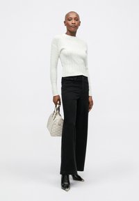 Suéter branco canelado combinado com jeans pretos de cintura alta. A modelo segura uma mala cinza clara com padrão de logótipos e usa botins pretos de salto.