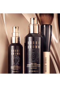 Svarte glassflasker med gullakenter, merket "Bobbi Brown Intensive Serum Foundation," ved siden av en svart og trefundamentbørste.