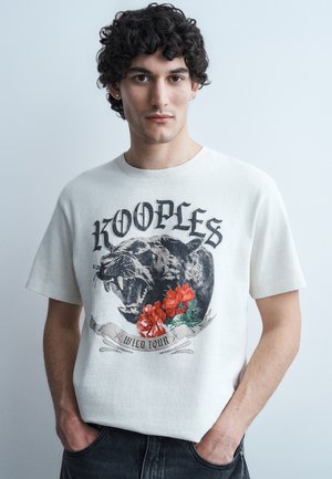 Νεαρός άνδρας με σγουρά μαλλιά που φοράει λευκό μπλουζάκι με σχεδιασμό λιονταριού που ουρλιάζει, κόκκινα λουλούδια και το κείμενο «KOOPLES WILD TOUR», με τα χέρια στις τσέπες.